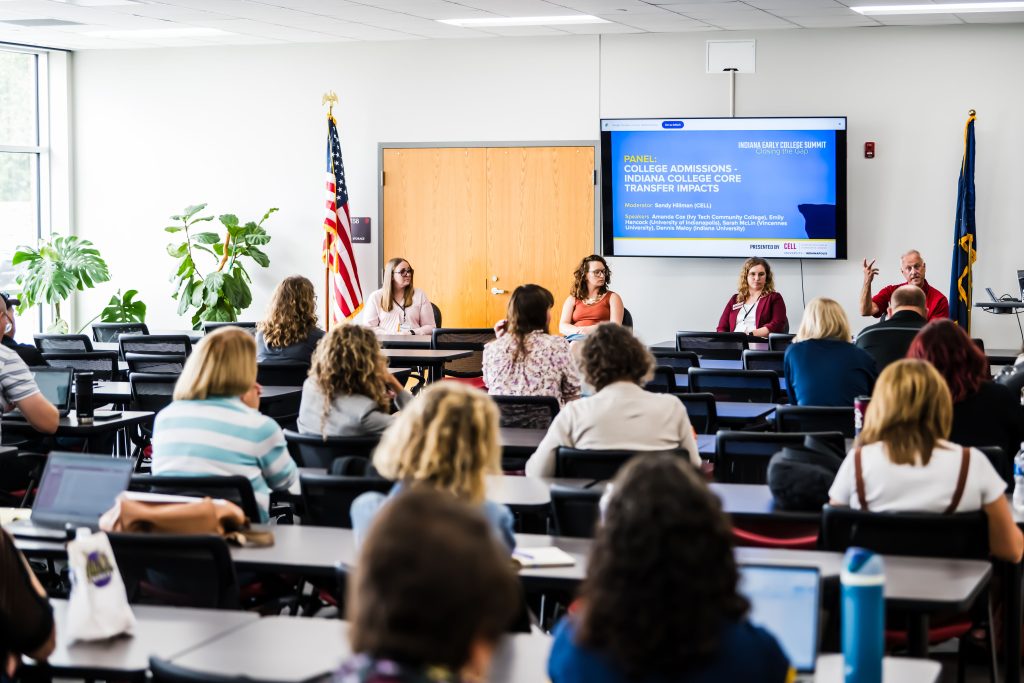 Room of people at desks with four panelists in front of presentation at front of room